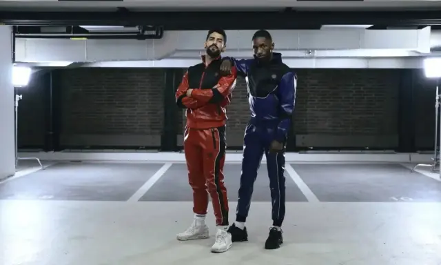 Two gay men in matching red and blue sportswear pose in a parking garage.