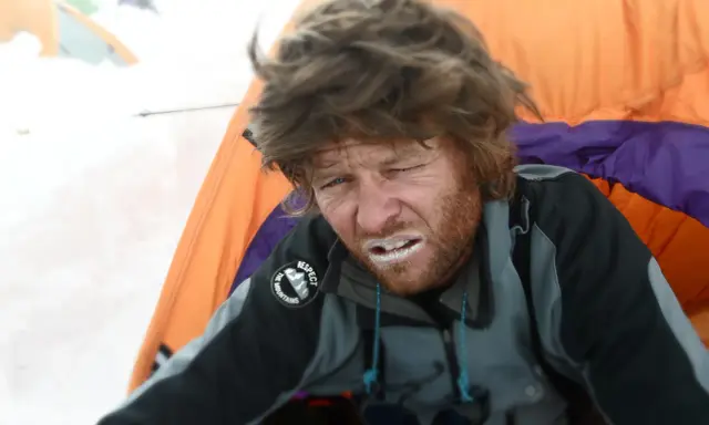 Zmęczony alpinista z białym nalotem na ustach, prawdopodobnie objawy choroby wysokościowej. W tle namioty.