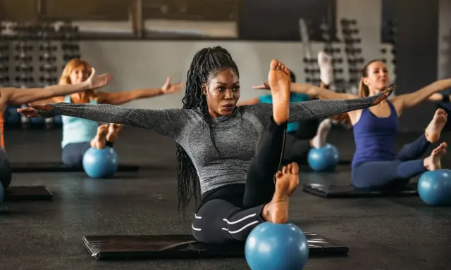 Frauen in einem Kurs, der Pilates Krafttraining macht, sitzen auf Matten mit kleinen blauen Bällen unter ihren Füßen.