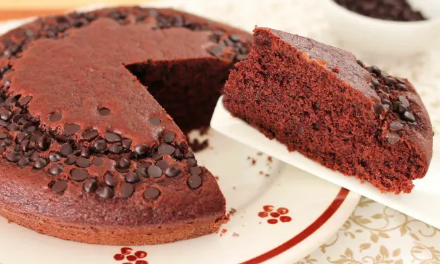 Una fetta di torta al cioccolato morbida, cosparsa di gocce di cioccolato, viene servita da una tortiera intera.