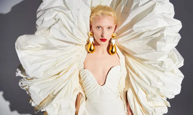 A model wears a dramatic white gown with a voluminous, ruffled cape, showcasing Daniel Roseberry's avant-garde design and striking gold teardrop earrings.