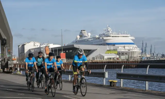 Fünf Radfahrer in blauen Trikots trainieren für einen Mini-Triathlon am Hafen, ein großes Kreuzfahrtschiff im Hintergrund.