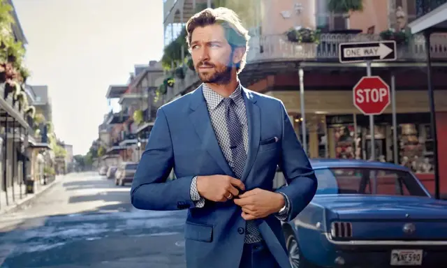 Man in a sharp blue suit, ready for a business cocktail, stands on a street with a vintage car and classic architecture.