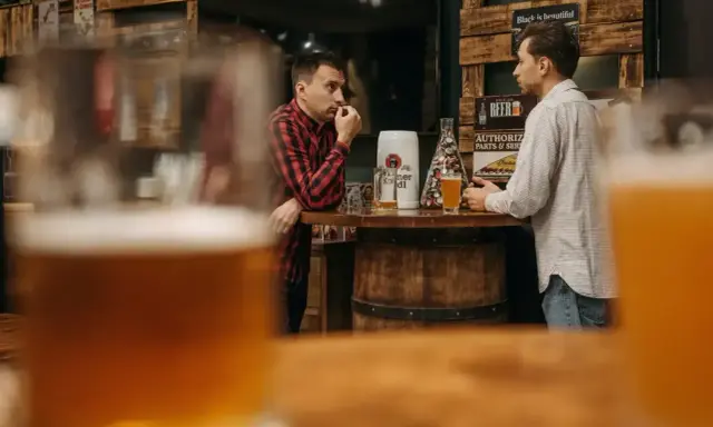 Dwóch mężczyzn rozmawia przy barze, zastanawiając się, ile gram alkoholu ma piwo. Na blacie stoją kufle i piwo.