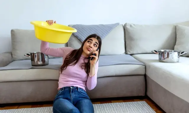 Mujer preocupada llama por teléfono mientras sostiene un barreño amarillo para recoger agua de las goteras en casa. Ollas esperan en el sofá.