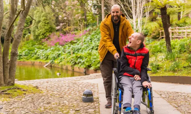 Mężczyzna pcha wózek z uśmiechniętym chłopcem. W tle piękny park z jeziorkiem. Pokazuje, że niepełnosprawność sprzężona nie ogranicza radości życia.