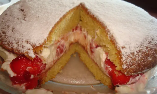 Puszysty biszkopt przełożony kremem i truskawkami, posypany cukrem pudrem. Klasyczny victoria sponge cake.