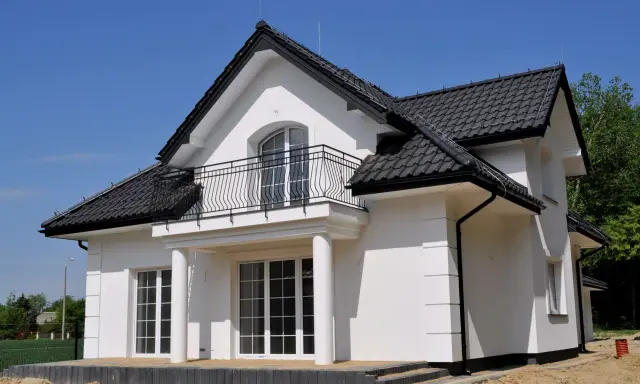 Nowy dom z białym gipsem na zewnątrz, czarnym dachem i balkonem. Wokół plac budowy z piaskiem.