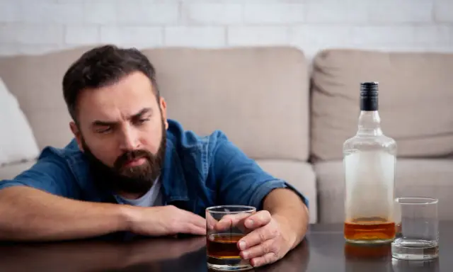 Mężczyzna z zarostem patrzy na szklankę z alkoholem, widoczne objawy uzależnienia. Obok pusta butelka i kolejna szklanka.