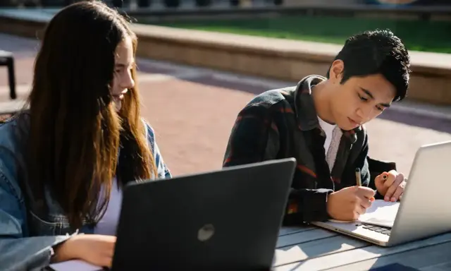 Dwoje studentów korzysta z laptopów na zewnątrz, przygotowując się do zajęć. To idealny przykład elastyczności, jaką oferują studia hybrydowe.