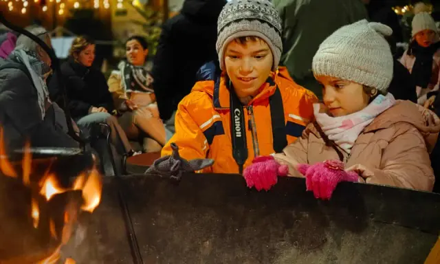 Niños disfrutan de una fogata en Italia, con luces festivas de fondo.