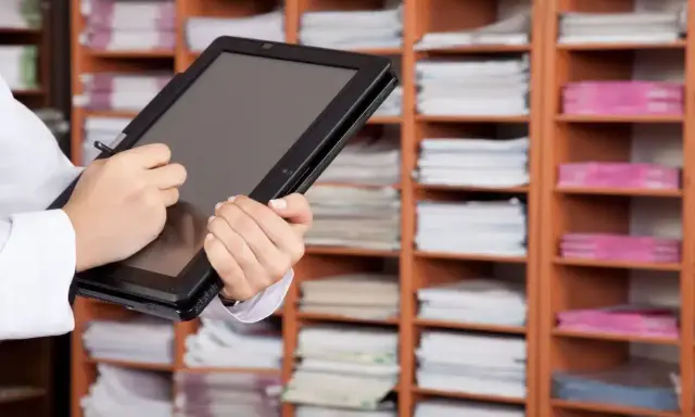 Une personne utilise une tablette pour gérer des documents, illustrant l'archivage électronique, une obligation légale. Les étagères remplies de dossiers rappellent l'importance de la numérisation.