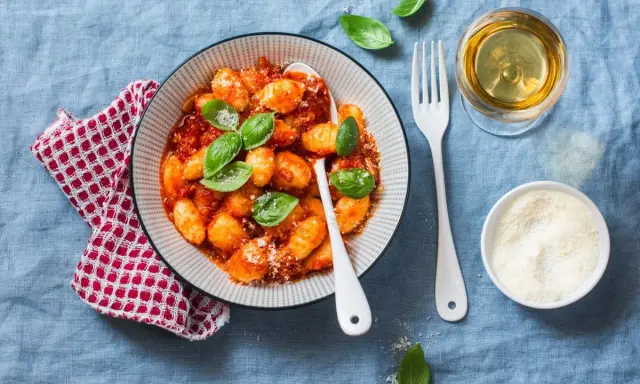 Pyszne kluski gnocchi w sosie pomidorowym, udekorowane bazylią. Obok kieliszek białego wina i miseczka parmezanu.