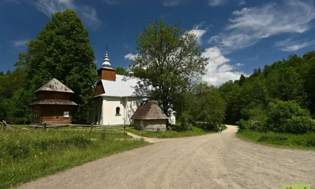 Cerkiew w Łopience, drewniana chata i kamienna przybudówka na tle zieleni i błękitnego nieba z chmurami.