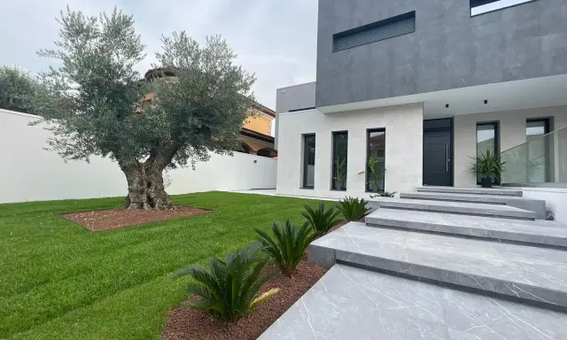 Moderna casa con fachadas color gris y blanco, escaleras de mármol y un olivo centenario en el jardín.