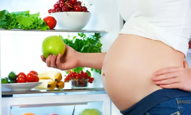 Gravid kvinna tar ett grönt äpple ur kylen fylld med färsk frukt och grönsaker. Hon njuter av hälsosam mat under sin soja graviditet.
