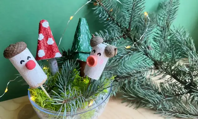 Uroczy stroik DIY z korkowych ludzików, grzybków i choinek w szklanej misie, ozdobiony gałązkami świerku i lampkami.