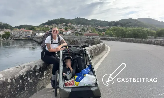 Frau mit Rucksack und Kinderwagen auf einer Brücke, bereit für eine abc tour. Im Hintergrund eine Stadt und Berge.