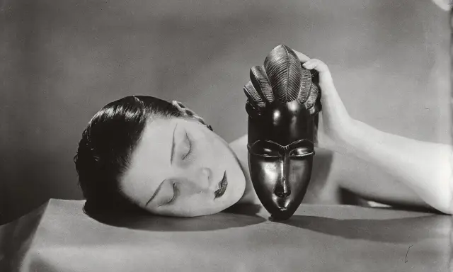 Foto Man Ray: volto femminile addormentato accanto a una maschera africana tenuta da una mano.