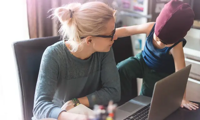 Une mère travaille sur son ordinateur portable, son enfant curieux s'approche. Un moment de vie quotidienne illustrant l'équilibre digital.