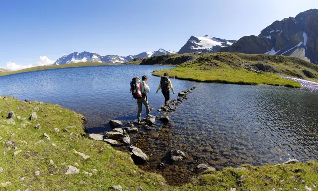 Due escursionisti attraversano il lago di Ceresole su un sentiero di pietre, con montagne innevate sullo sfondo.
