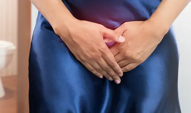 Kobieta w granatowej sukience z rękami na podbrzuszu, w tle toaleta. Problem z nietrzymaniem moczu? Czas na wizytę u lekarza.