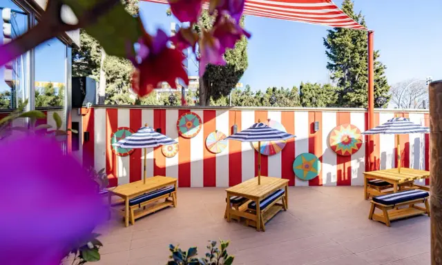 Terraza de restaurantes con zona infantil en Madrid, decorada con rayas rojas y blancas y coloridos adornos circulares. Mesas de madera con sombrillas a rayas invitan a disfrutar.