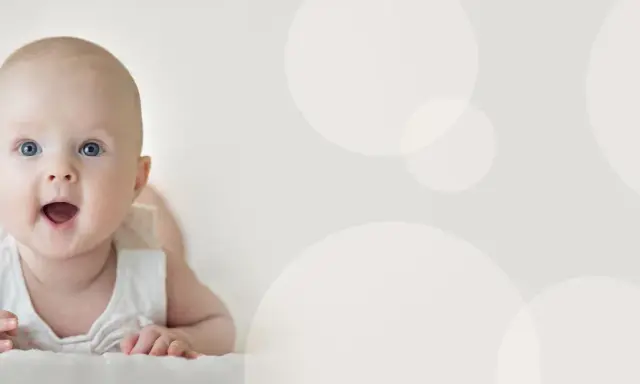 Uśmiechnięte niemowlę 4 miesiące leży na brzuchu, z otwartymi ustami i szeroko otwartymi oczami, patrząc wprost na widza.