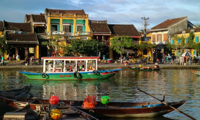 Båtar flyter på floden i Hoi An, Vietnam, med färgglada hus och folk som promenerar längs kajen.