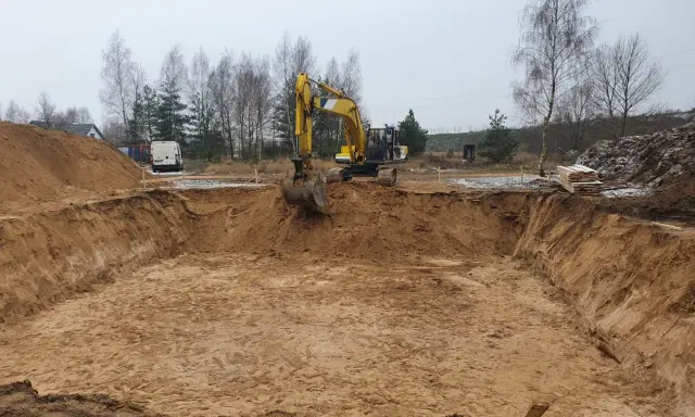 Żółty koparka wykonuje wykopy pod fundament. W tle widać stosy piasku i drzewa.