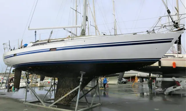 Un grand voilier blanc et bleu est sur des chandelles, son flanc couvert de bernacles. Un homme nettoie la coque d'un autre bateau à proximité.