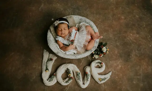 Dolce foto neonato 1 mese dorme in una cesta, circondato da fiori e dalla parola "love" creata con tessuto.