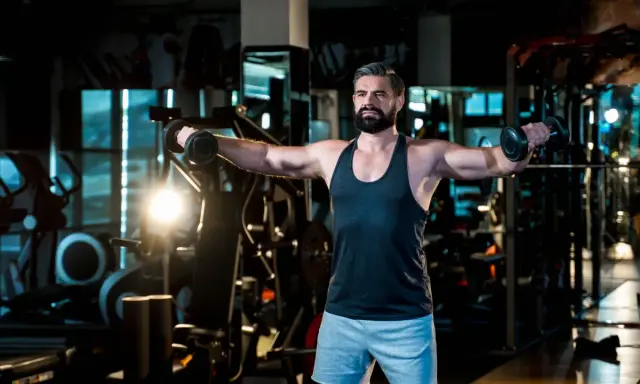 Mann mit Bart trainiert im Fitnessstudio, macht seitheben sitzend mit Hanteln.