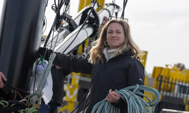 Élodie Bonafous, souriante, tient une corde bleue sur un bateau, entourée de cordages et de structures métalliques.