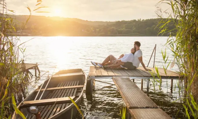 Para, gdzie na Mazury we dwoje? Para siedzi na pomoście, łódka przy brzegu, zachód słońca.