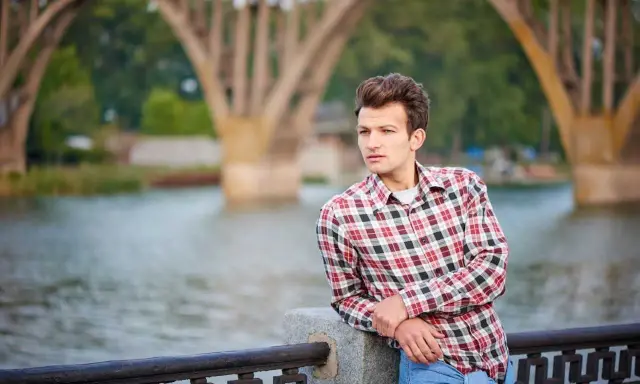 Młody mężczyzna w kratowanej koszuli i dżinsach opiera się o balustradę nad rzeką. W tle widać most.