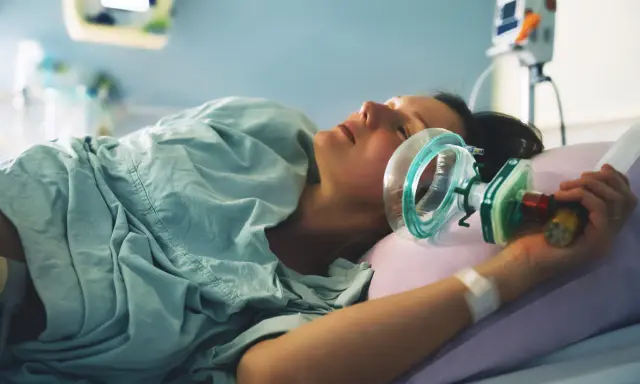 Kobieta w szpitalnym łóżku, z maską gazu rozweselającego przy twarzy, przygotowuje się do porodu.