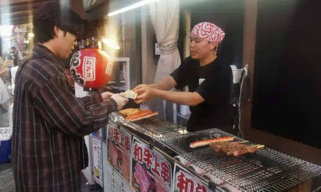 Un cliente paga por comida callejera japonesa: brochetas de carne y surimi a la parrilla en un puesto.