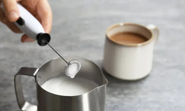 Hand hält Milchaufschäumer über Metallkännchen mit Milch. Perfekt für leckere Milchschaum Rezepte.