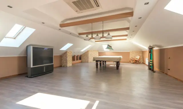 Vaste pièce sous les combles aménageables, avec table de billard, TV, fauteuil à bascule et étagères. Idéal pour un espace de loisirs.