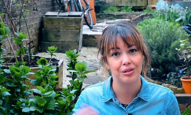 Mujer sonriente en un jardín con plantas verdes y macetas.