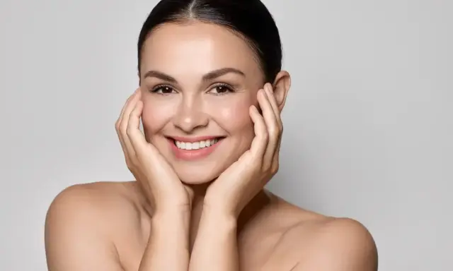 Uśmiechnięta kobieta z idealną, lśniącą cerą typu "glass skin", delikatnie dotykająca policzków dłońmi.