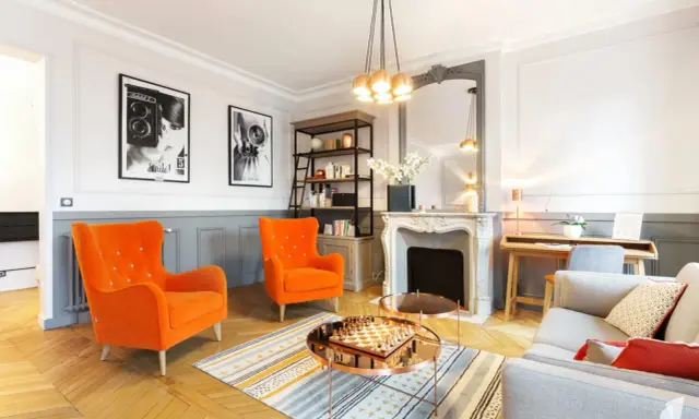 Salon aux fauteuils orange vif, cheminée ornée, et un jeu d'échecs sur table basse, évoquant un mouvement baroque moderne.