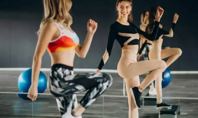 Dwie kobiety ćwiczą na stepach. Jedna w kolorowym topie i wzorzystych legginsach, druga w czarnym body i beżowych legginsach. TBC co to znaczy?