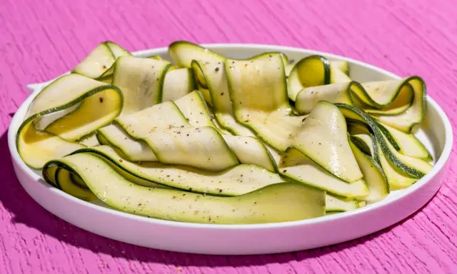 Fettine sottili di zucchine marinate crude, condite con pepe nero, su un piatto bianco, su un tavolo rosa acceso.