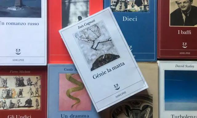 Libri Adelphi in primo piano, tra cui "Génie la matta" di Inès Cagnati, con un albero spoglio su sfondo grigio.