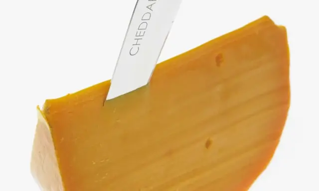 Ein Messer schneidet ein Stück leuchtend orangefarbenen Cheddar-Käse. Was ist Cheddar? Ein köstlicher Hartkäse.