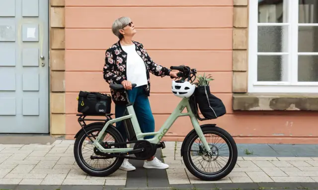 Ältere Dame mit QIO Bike, positive Erfahrungen mit dem E-Bike, das mit einem Helm und einer Tasche beladen ist.
