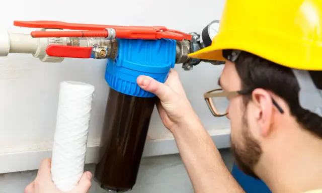 Un homme en casque jaune effectue la régénération manuelle d'un adoucisseur d'eau, tenant un nouveau filtre.