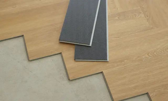 Panele winylowe w trakcie układania na betonowej podłodze. Dwa szare panele leżą na drewnianych deskach, pokazując, jak układać panele winylowe.